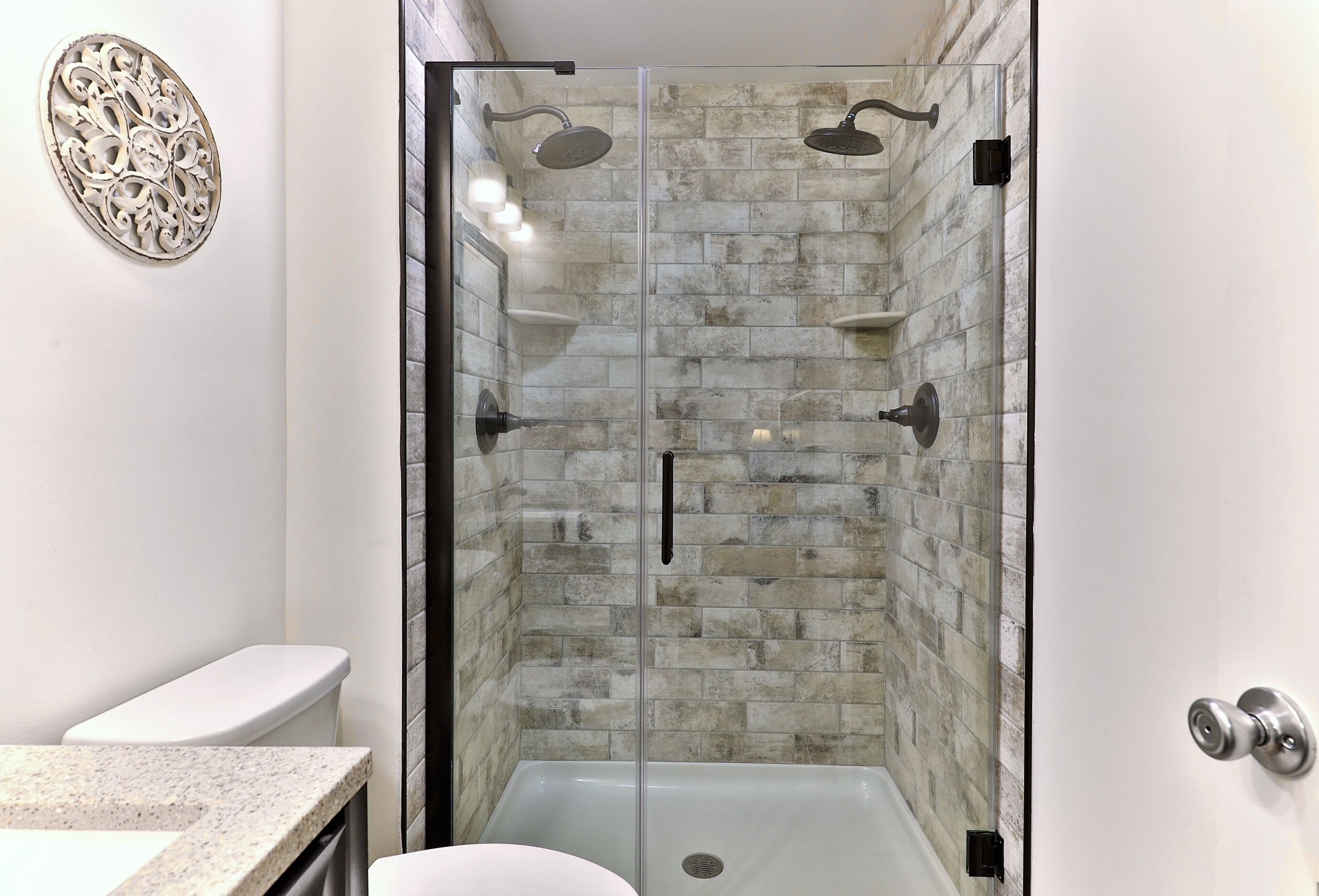 A glass-enclosed shower with a brick wall, a white toilet, and a granite countertop.