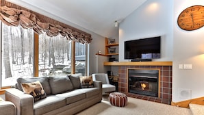Una sala de estar acogedora con chimenea, un ventanal con vistas a un bosque nevado, un sofá gris con cojines decorativos y un televisor de pantalla plana sobre la chimenea.