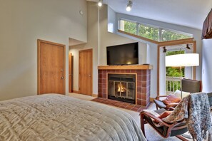 A bedroom with a large bed, a fireplace, a television, and a view of greenery through a window.