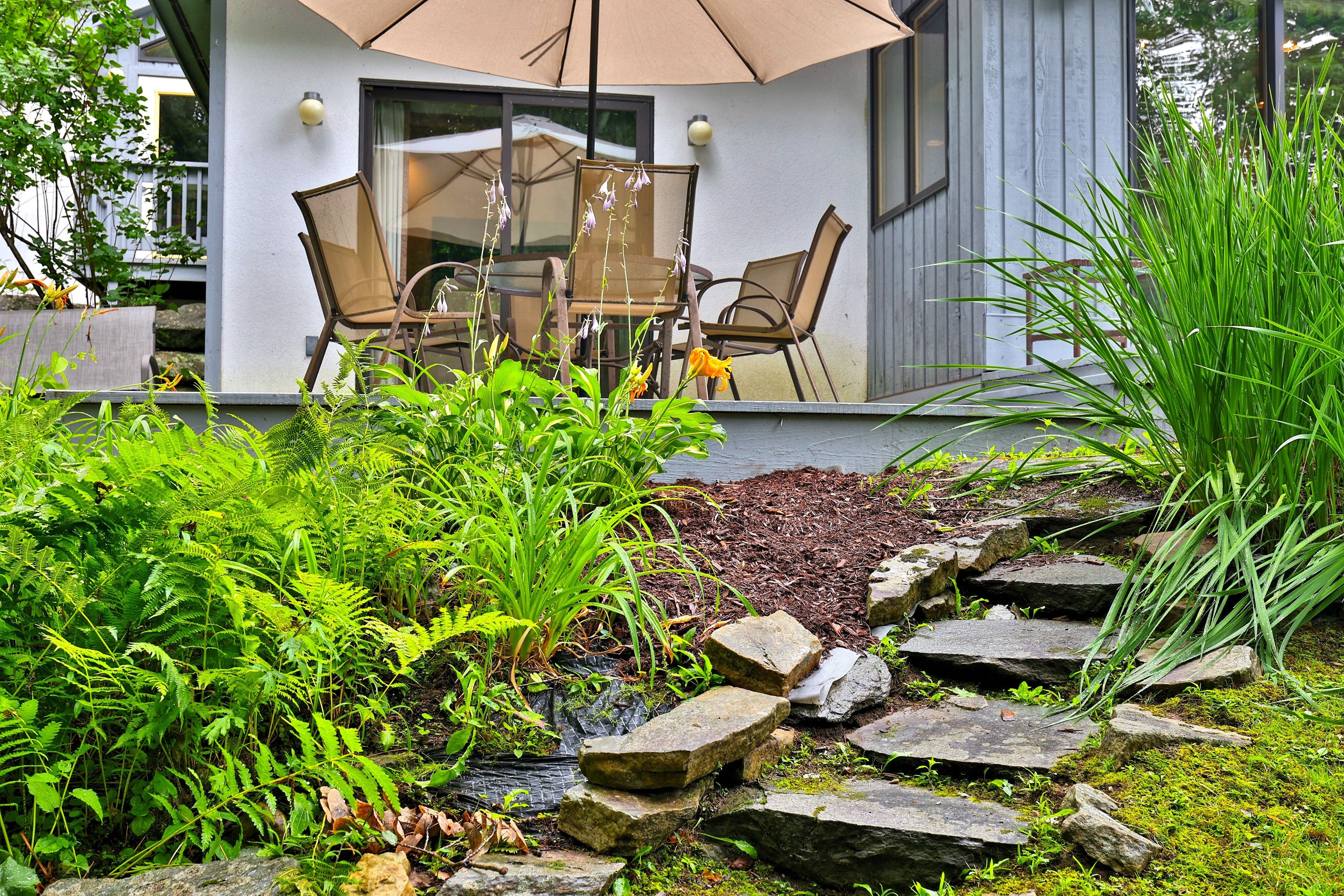 A modern outdoor seating area with a table and chairs, surrounded by lush greenery and a stone pathway.
