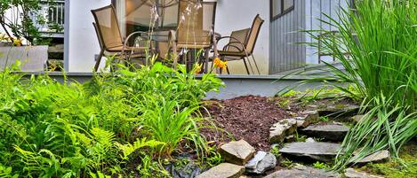 A modern outdoor seating area with a table and chairs, surrounded by lush greenery and a stone pathway.
