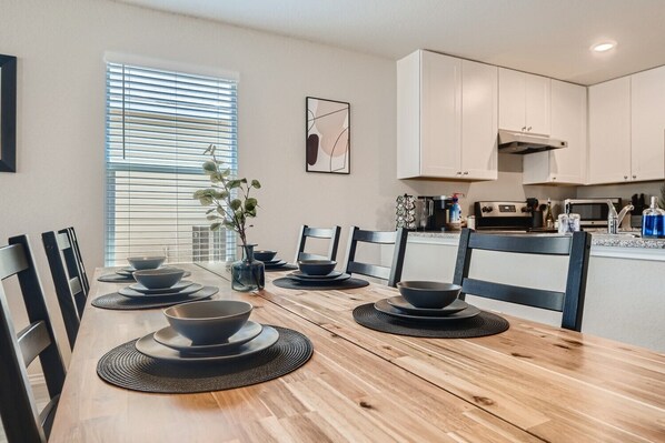 A dining area with a wooden table set for six, surrounded by chairs. A window with blinds is visible, and a kitchen area with white cabinets and appliances is in the background.