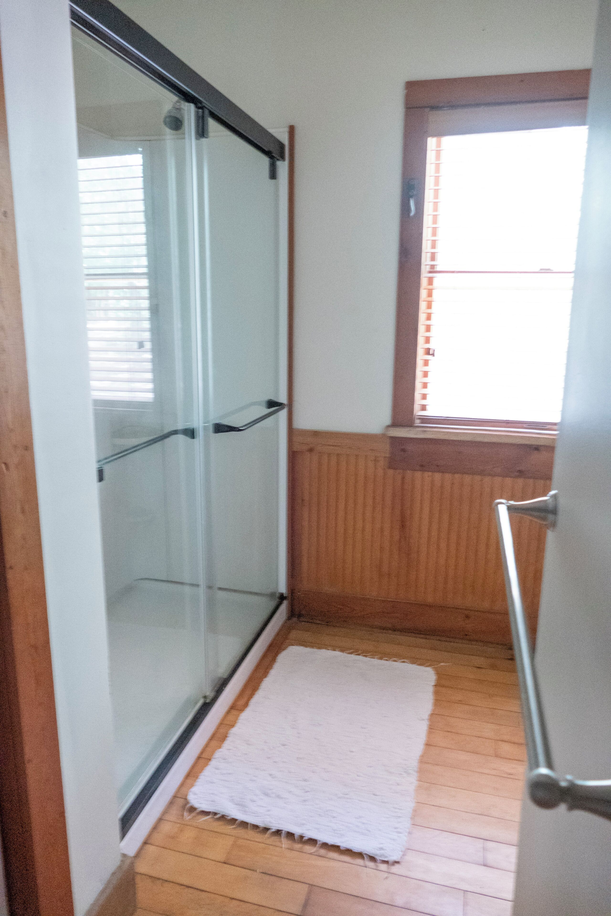A bathroom with a glass shower door, wooden flooring, a white rug, and a window with blinds.
