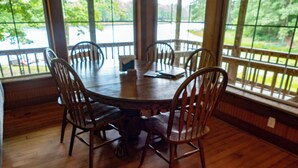 A round wooden dining table with six chairs, large windows with a view of greenery, and a wooden floor.