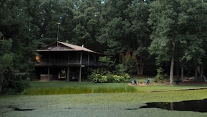 A two-story wooden cabin with a balcony, surrounded by dense forest and a pond.
