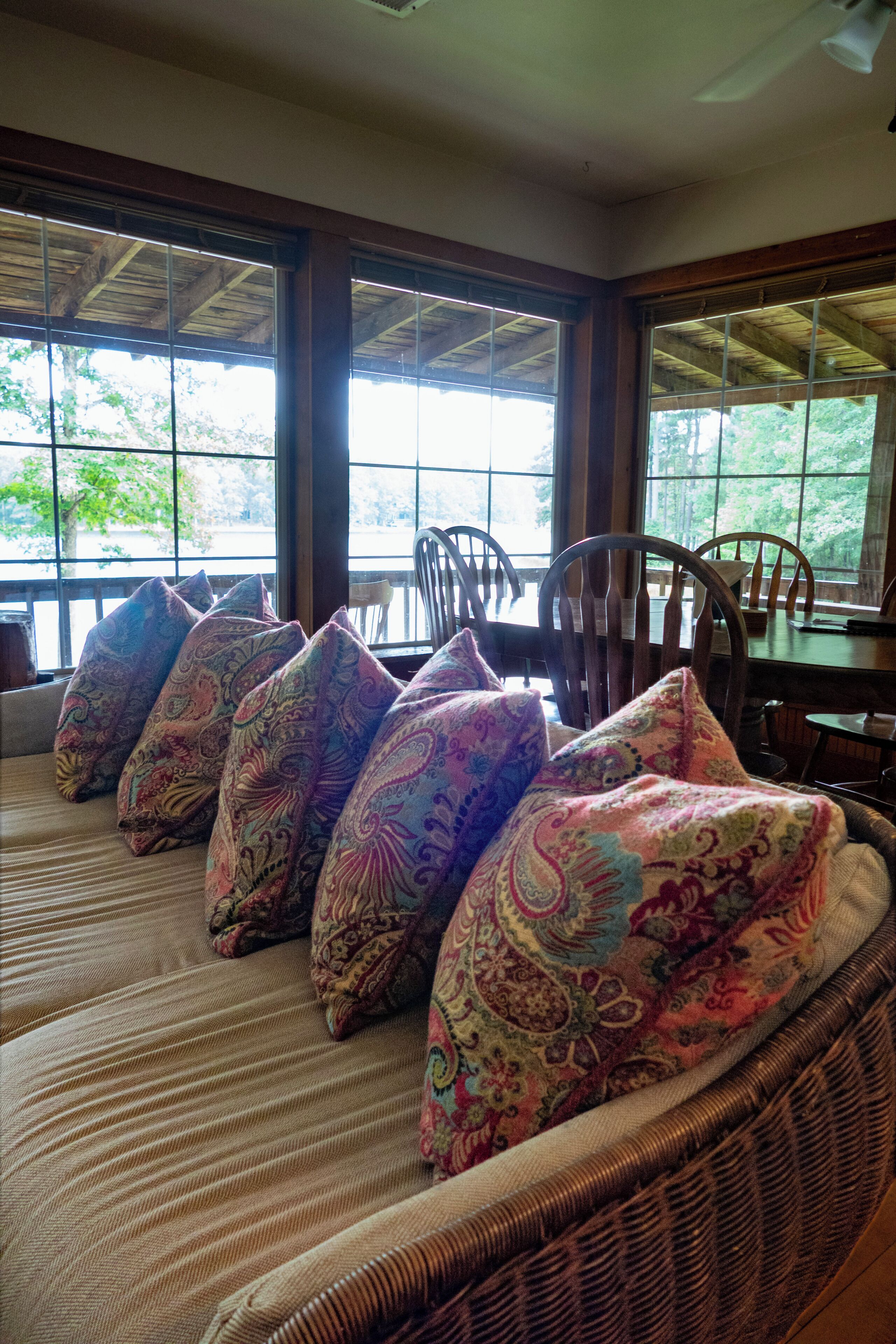 A room with a large window, a wicker sofa with patterned cushions, and a wooden table.