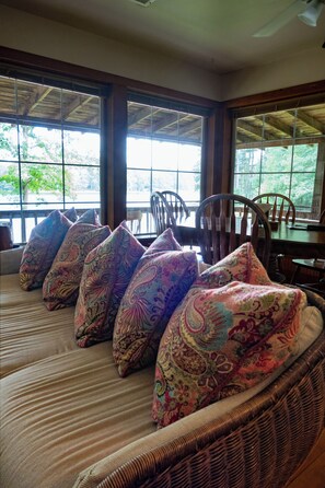 A room with a large window, a wicker sofa with patterned cushions, and a wooden table.