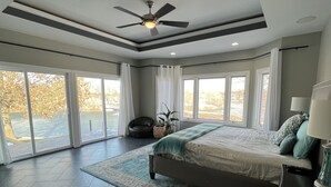 Une chambre dotée d’un grand lit, d’un ventilateur de plafond et offrant une vue sur l’extérieur grâce à de grandes fenêtres.