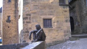 Une statue de pierre représentant une figure assise, placée sur une corniche devant un bâtiment aux murs de pierre et doté d’une fenêtre.