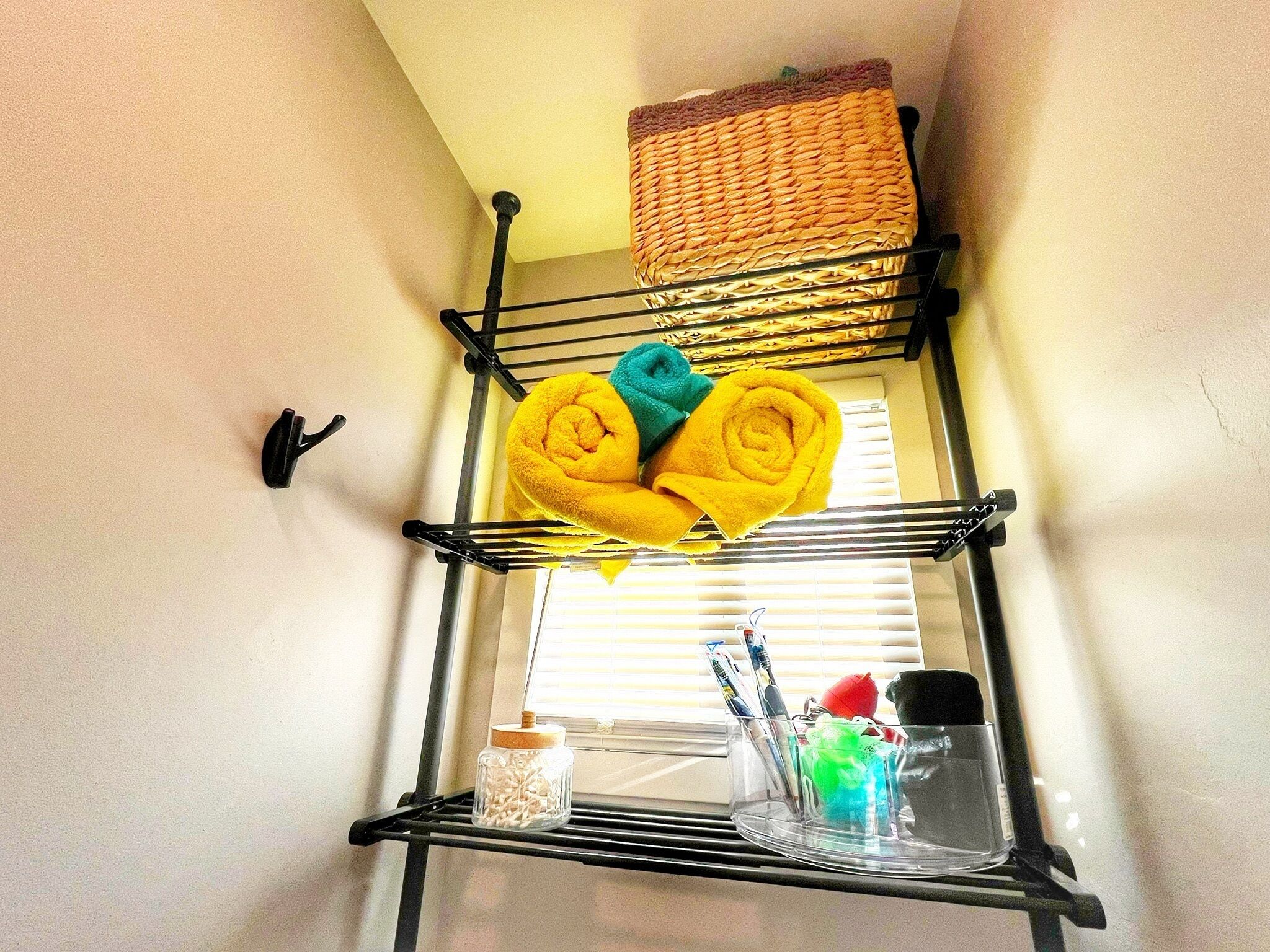 A corner with a black metal shelf unit holding rolled towels and a basket.