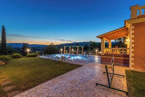A pool area with lounge chairs and a pergola, surrounded by a stone patio and a grassy lawn.