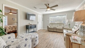 A spacious living room with a beige sofa, a recliner, and a glass coffee table. There is a ceiling fan, a TV mounted on the wall, and a window with blinds. - Charming Gulfport Home ~ 2 Mi to Marina (Gulfport)