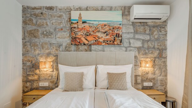 A bedroom with a stone wall, a bed with white linens, two bedside tables with lamps, and a painting of a coastal town. - InMotion Luxury Rooms (Split)