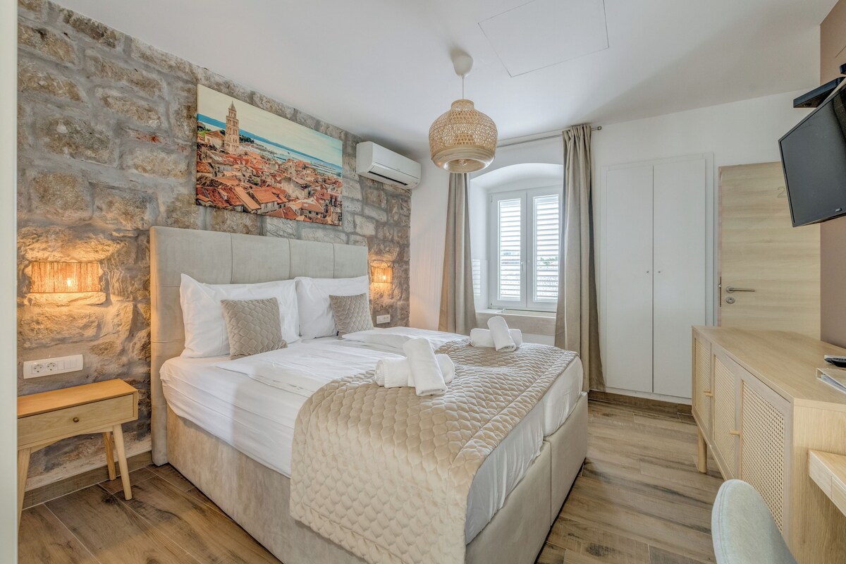 A bedroom with a stone wall, a bed with white bedding, a desk with a TV, and a painting of a cityscape.