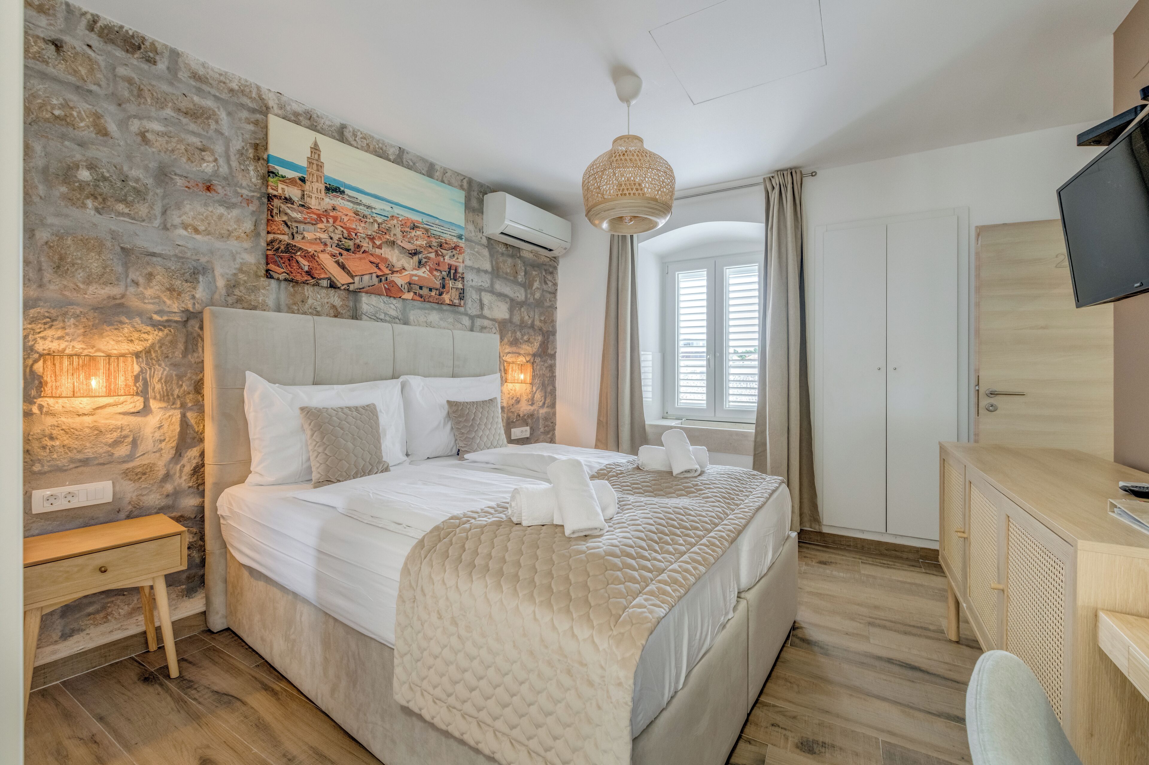 A bedroom with a stone wall, a bed with white bedding, a desk with a TV, and a painting of a cityscape.