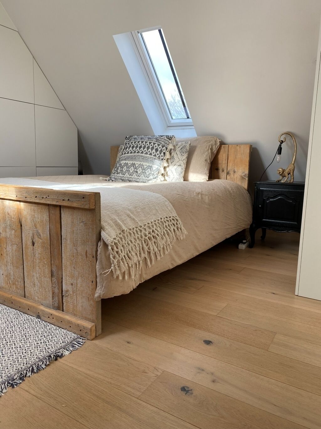 Une chambre à coucher aménagée dans un espace mansardé, avec un plafond en pente, un puits de lumière, un cadre de lit en bois, un tapis à motifs et une table de chevet avec une lampe.