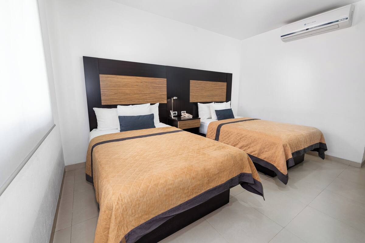 A hotel room with two beds, a bedside table with a lamp, and an air conditioning unit on the wall.