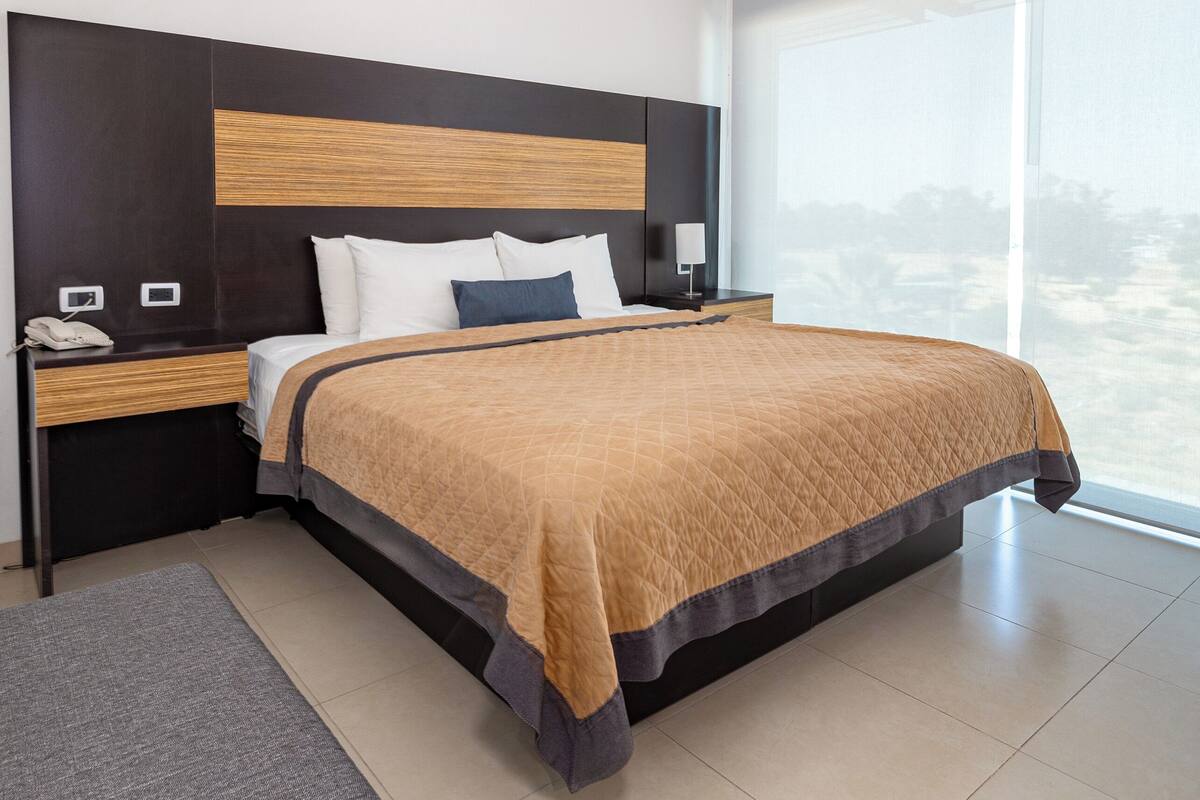 A modern hotel room with a large bed, a bedside table with a phone, a nightstand, and a bench.