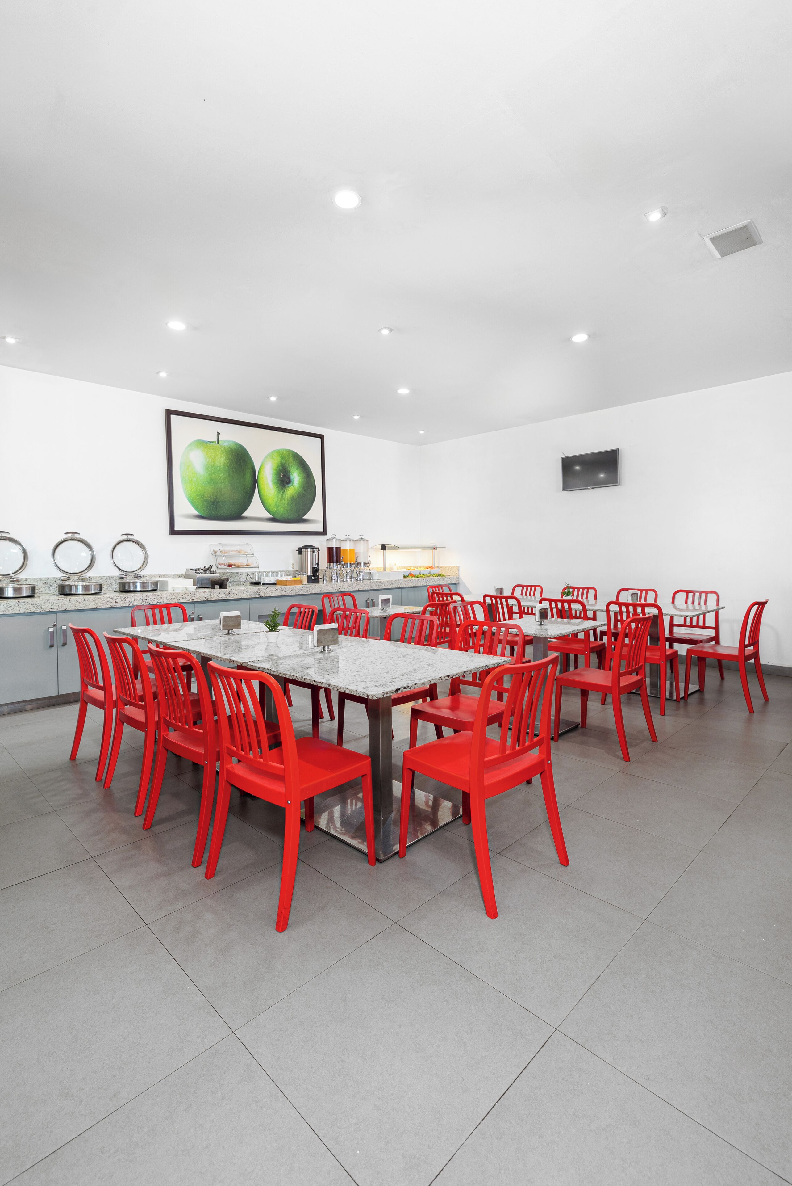 Un comedor moderno con una mesa larga y sillas rojas, una encimera de cocina con electrodomésticos, y un cuadro grande de una manzana verde en la pared.