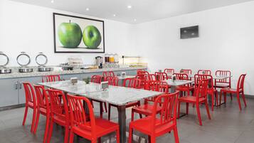 Un comedor moderno con una mesa larga y sillas rojas, una encimera de cocina con electrodomésticos, y un cuadro grande de una manzana verde en la pared.