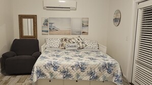 A bedroom with a bed, a chair, a wall-mounted air conditioner, and a beach-themed wall art. - Charming Beach-Themed Studio Apartment in Doerun, GA with Private Pool (Doerun)