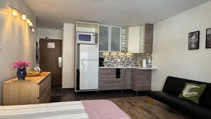 Un espace de vie compact comprenant un coin cuisine, un lit avec une couverture à rayures, un canapé noir avec un coussin vert et un vase rempli de fleurs roses posé sur une commode en bois.
