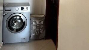 A laundry room with a front-loading washing machine, a colorful circular wall art piece, and a wooden door. - Shilo Space-Emerald Apt.Free WiFi & Modern Look. (Kampala)