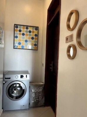 A laundry room with a front-loading washing machine, a colorful circular wall art piece, and a wooden door. - Shilo Space-Emerald Apt.Free WiFi & Modern Look. (Kampala)