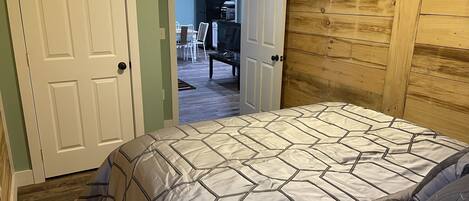 A bedroom with a bed, wooden panel walls, a door leading to a dining area, and a visible kitchen in the background.