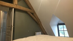 A room with a sloped ceiling, exposed wooden beams, a white door, and a window with a view of greenery. - 18th century character gîte in the VAL DE CHAMBORD at the gates of Chambord (Huisseau-sur-Cosson)