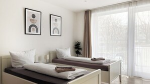 Et moderne hotelværelse med to senge, et sengebord, en potteplante og store vinduer med lette gardiner.