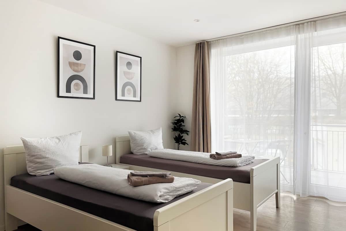 Une chambre d’hôtel moderne avec deux lits, une table de chevet, une plante en pot et de grandes fenêtres avec des rideaux légers.