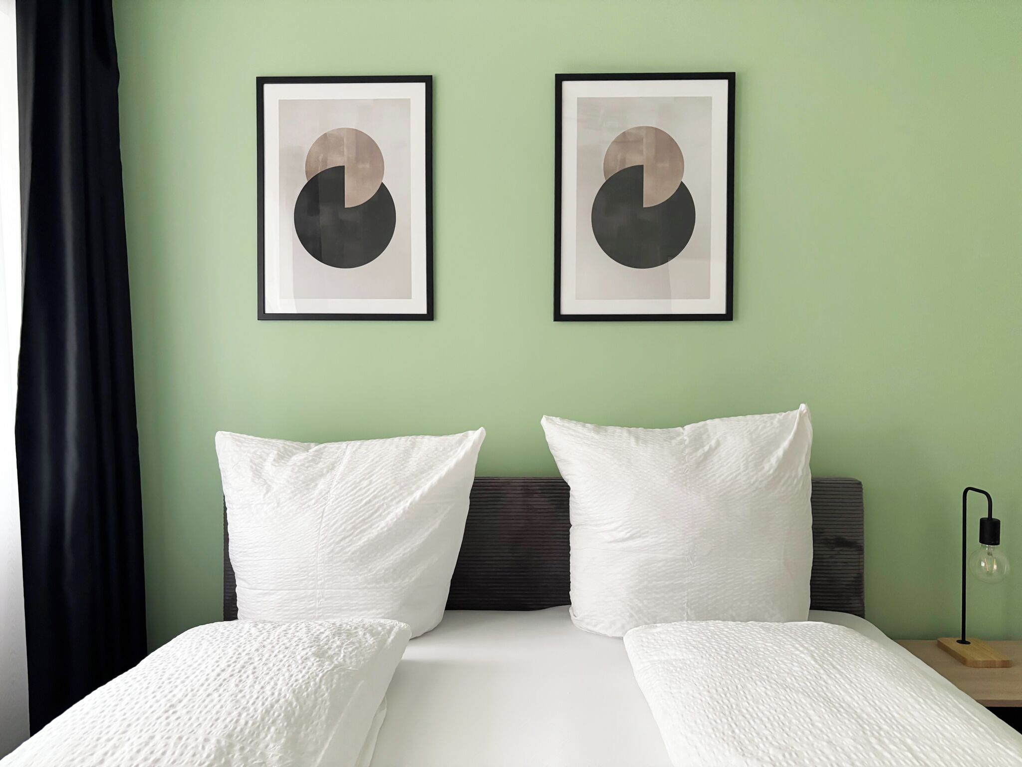 A hotel room with a bed, two pillows, a headboard, a nightstand with a lamp, and two framed abstract art pieces on the wall.