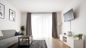 En moderne stue med en grå sofa, et sofabord, et fjernsyn på et stativ og en plante på et hvidt skab.