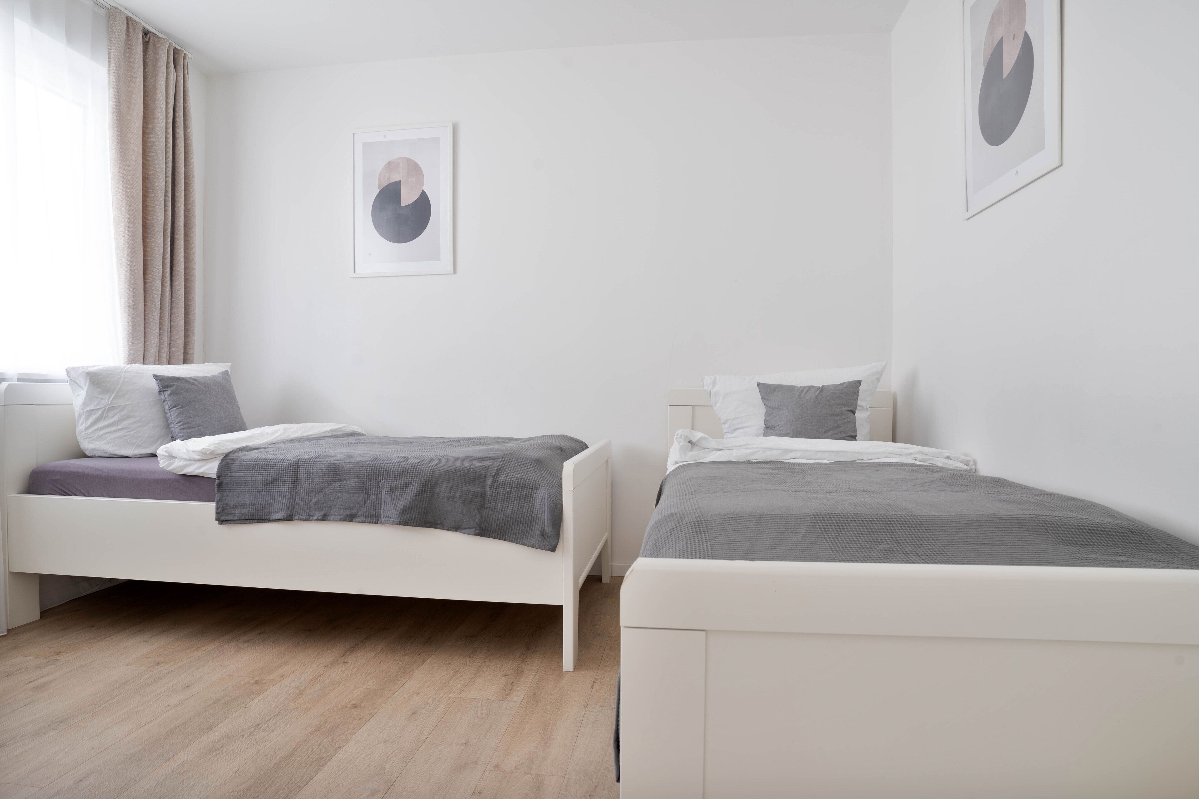 Een moderne slaapkamer met twee bedden, een raam met gordijnen en twee ingelijste kunstwerken aan de muur.