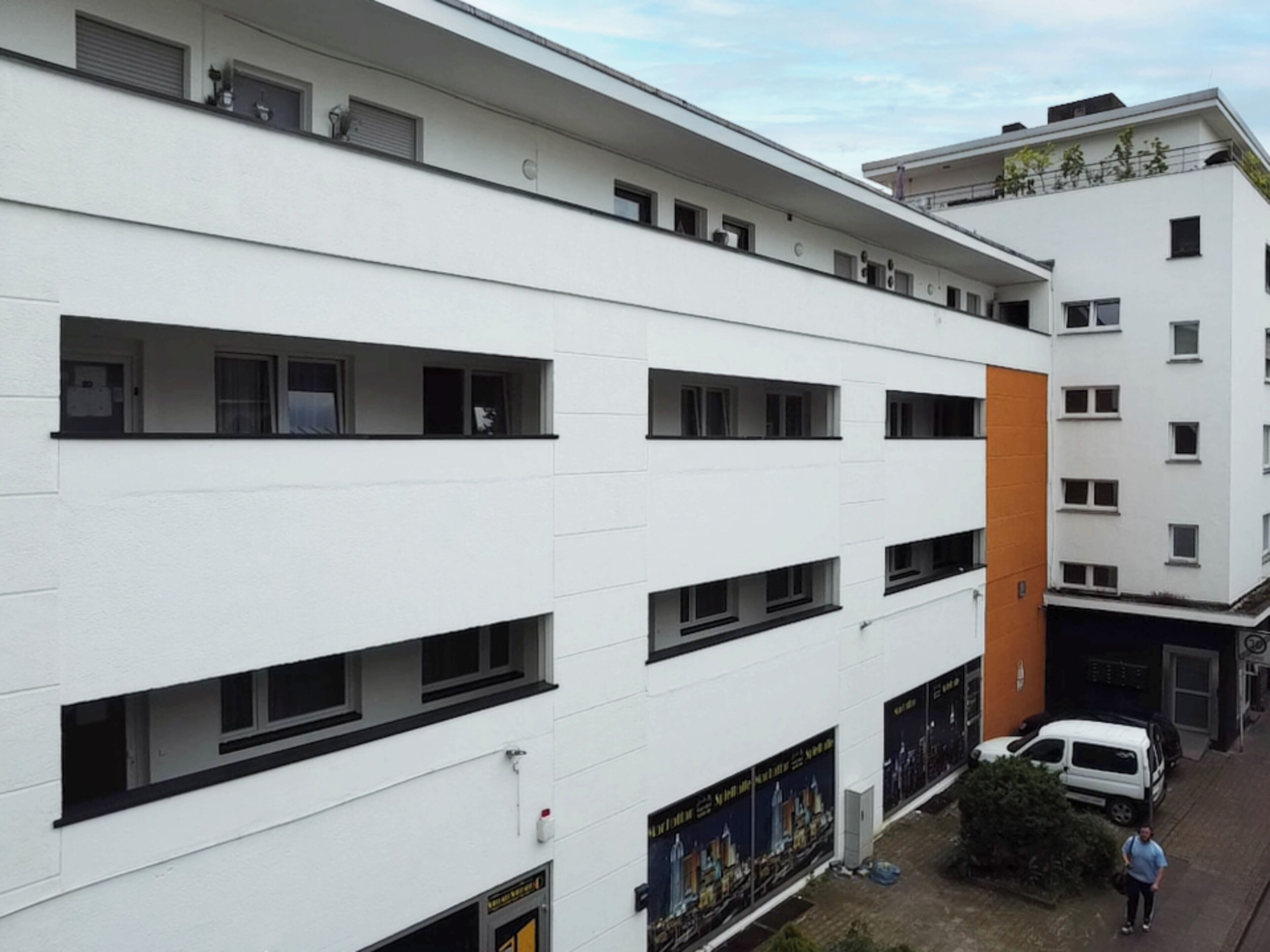 Ein mehrstöckiges Gebäude mit weißer Fassade, zahlreichen Fenstern und einem orangefarbenen Akzent.