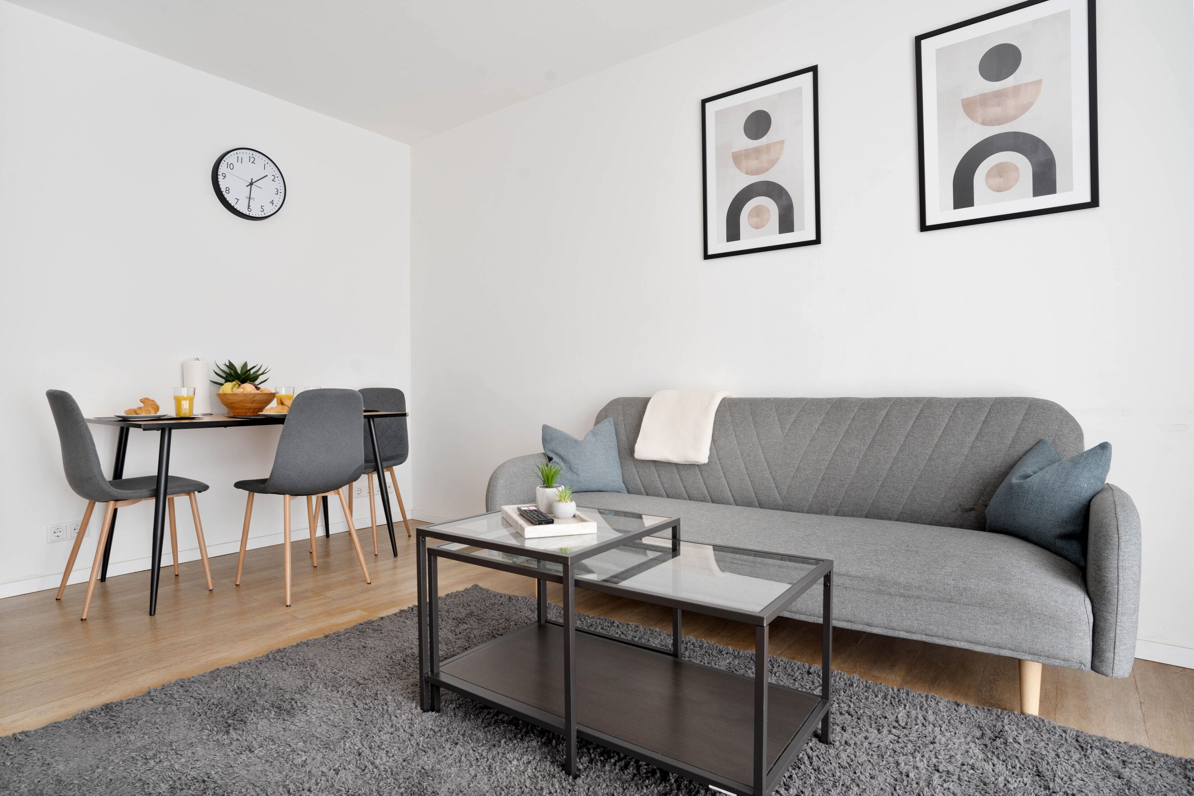 A modern living room with a grey sofa, a glass coffee table, and a dining area with a table and chairs.