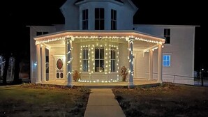 A large, white house with a wrap-around porch and a covered front porch area, featuring string lights and a number "291" sign.