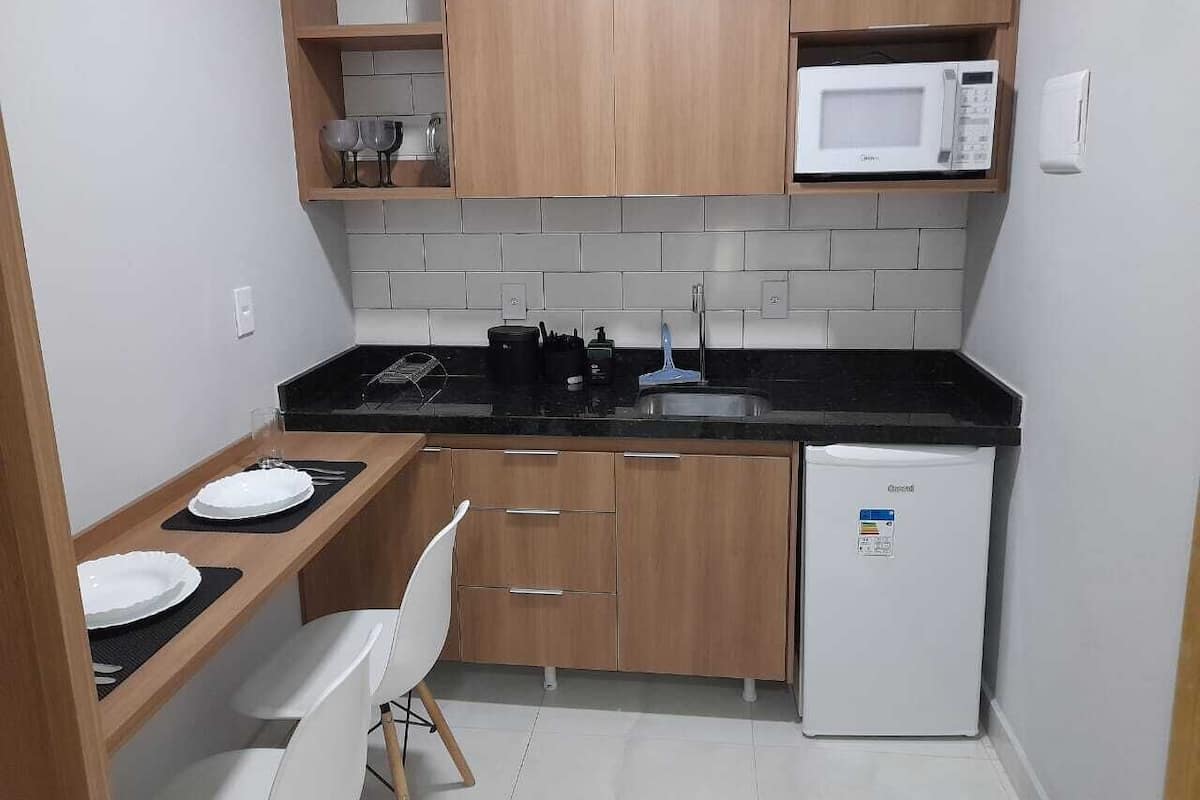 Una cocina compacta con armarios de madera, encimera negra, microondas, refrigerador y un pequeño comedor con una silla y platos.