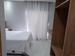 A hotel room with a bed, a wooden wardrobe, and a window with sheer curtains.