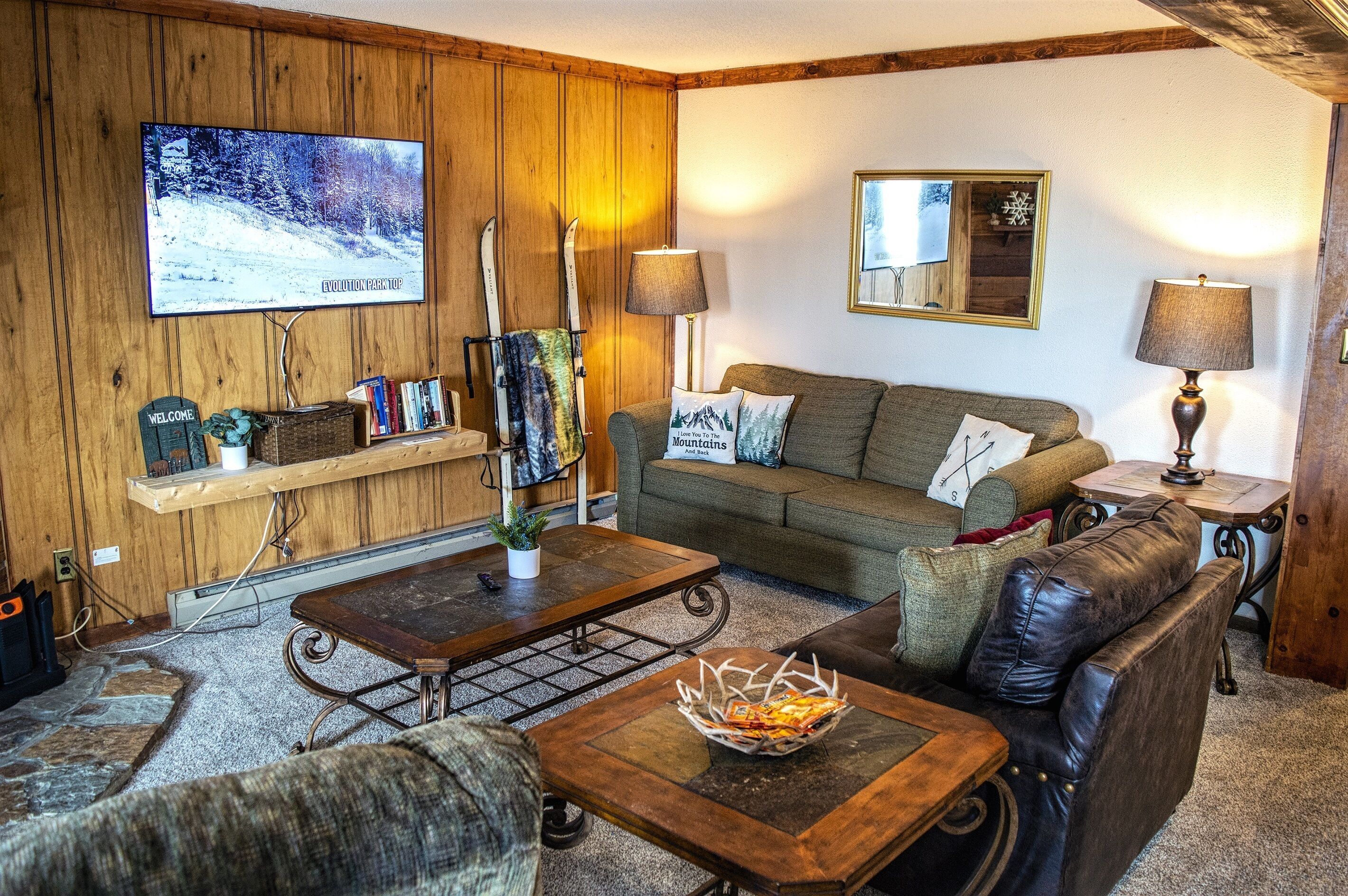 Een gezellige woonkamer met een houten wand, een televisiescherm waarop een besneeerd landschap te zien is, een bank met kussens, een salontafel en twee lampen.