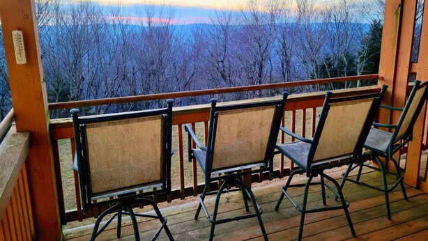 Eine Holzterrasse mit drei Stühlen, die einen Panoramablick auf eine bewaldete Landschaft bietet.