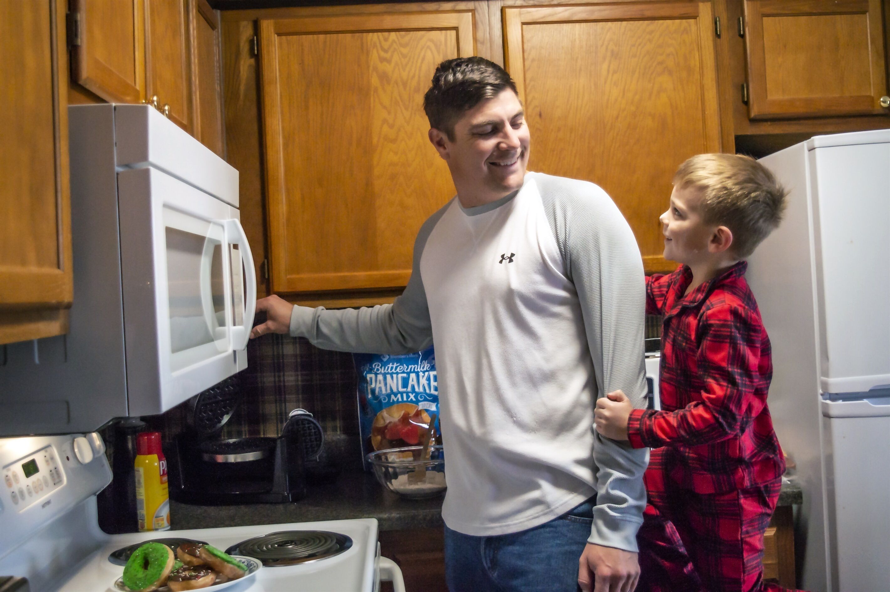 A kitchen with wooden cabinets, a microwave, a box of pancake mix, and a child standing next to an adult.