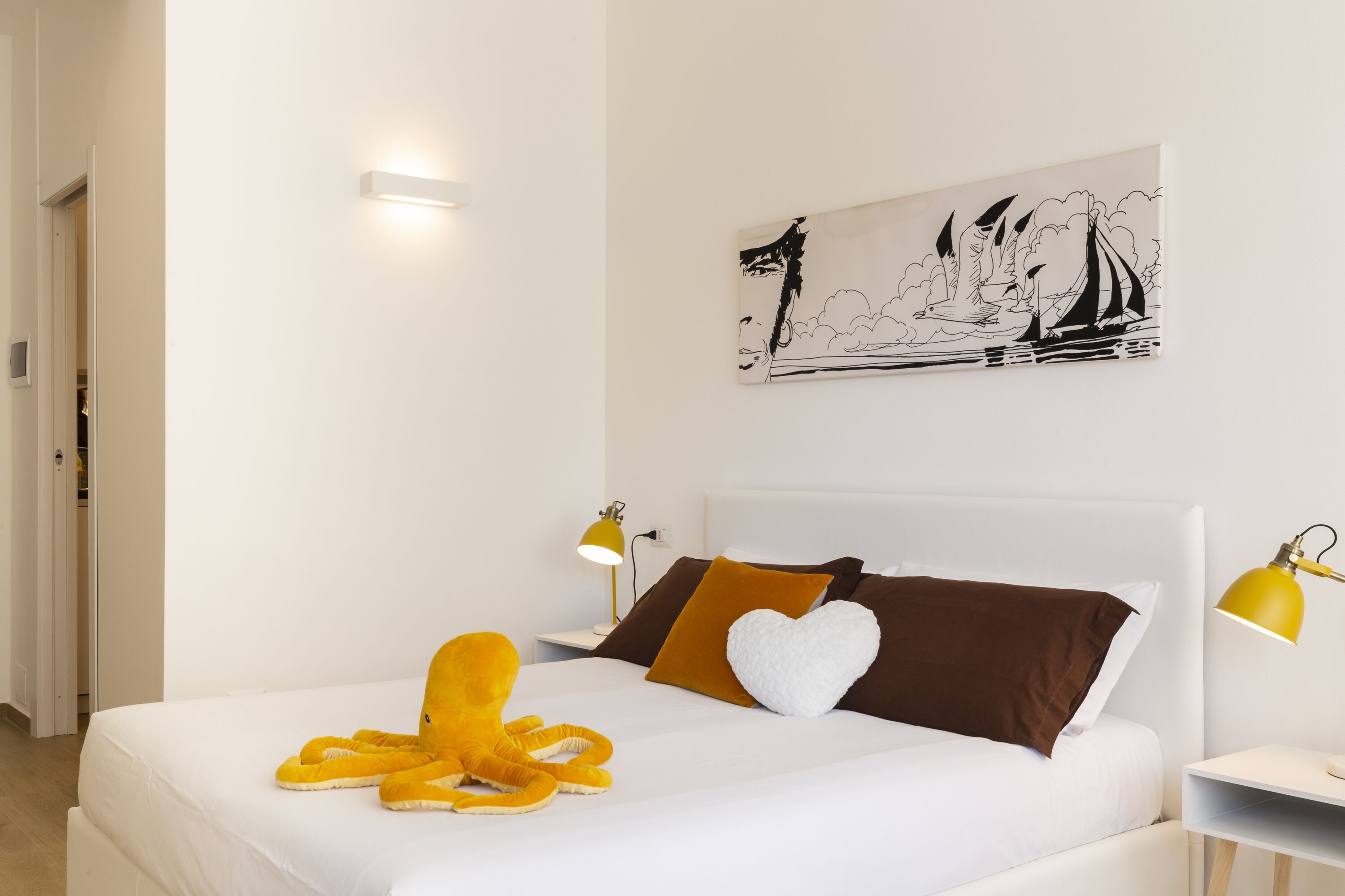 A neatly made bed with a white comforter, a brown pillow, a white heart-shaped pillow, and a yellow octopus plush toy. There is a black and white artwork on the wall depicting a sailboat and a figure. Two bedside lamps with yellow shades are on either side of the bed.