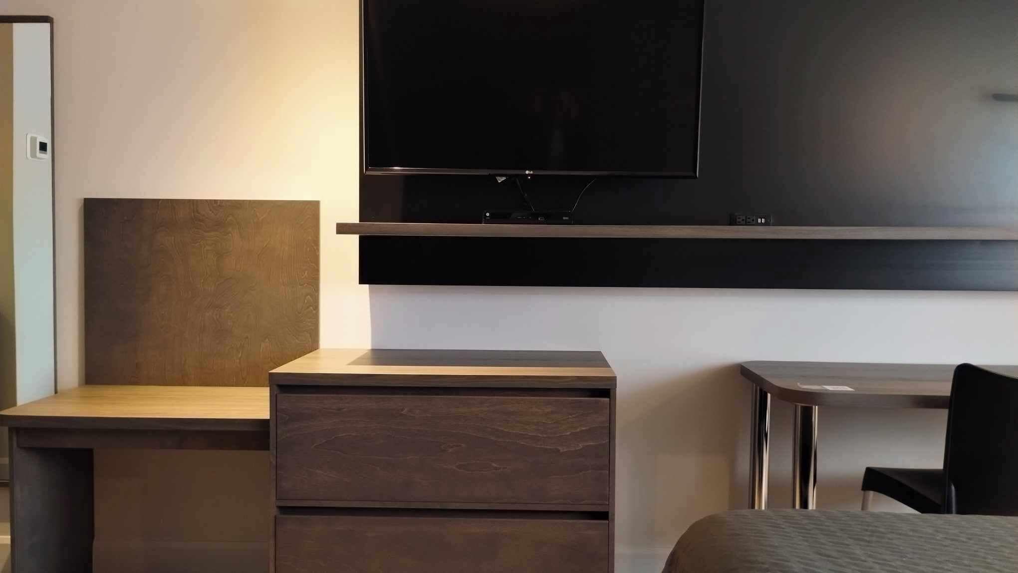 A modern hotel room with a flat-screen TV mounted on the wall, a wooden nightstand, a desk with a chair, and a bed.