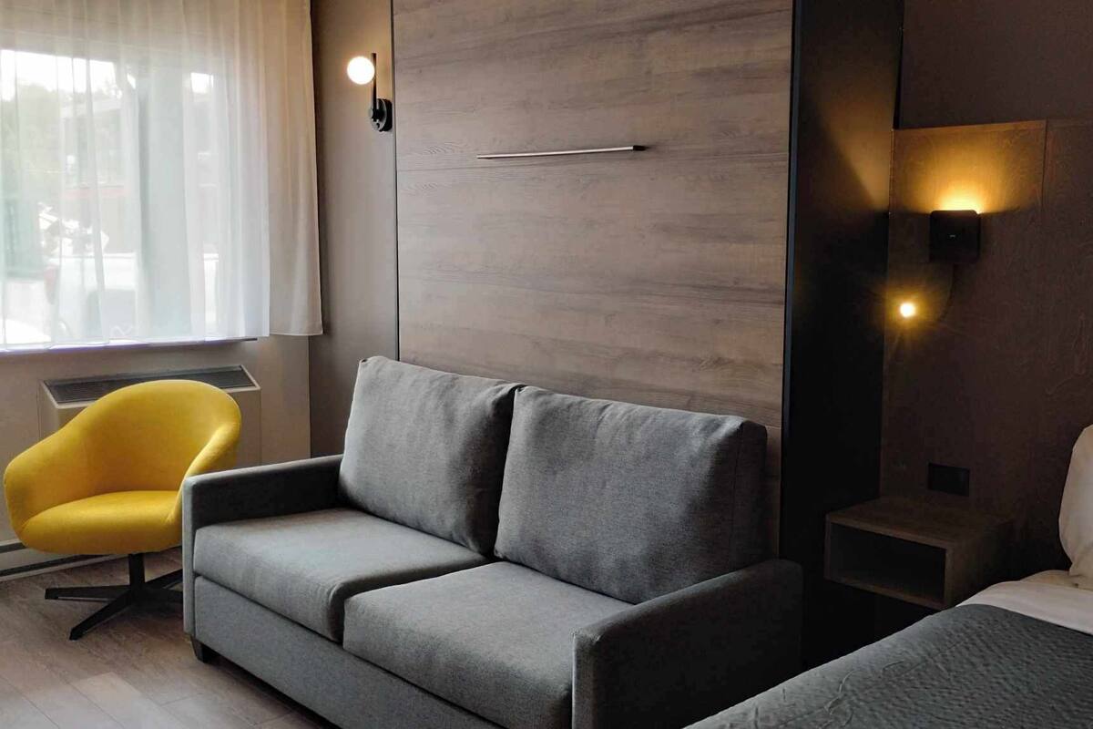 Une chambre d’hôtel moderne avec un canapé gris, un fauteuil jaune, un lit avec une literie blanche et deux lampes fixées au mur.