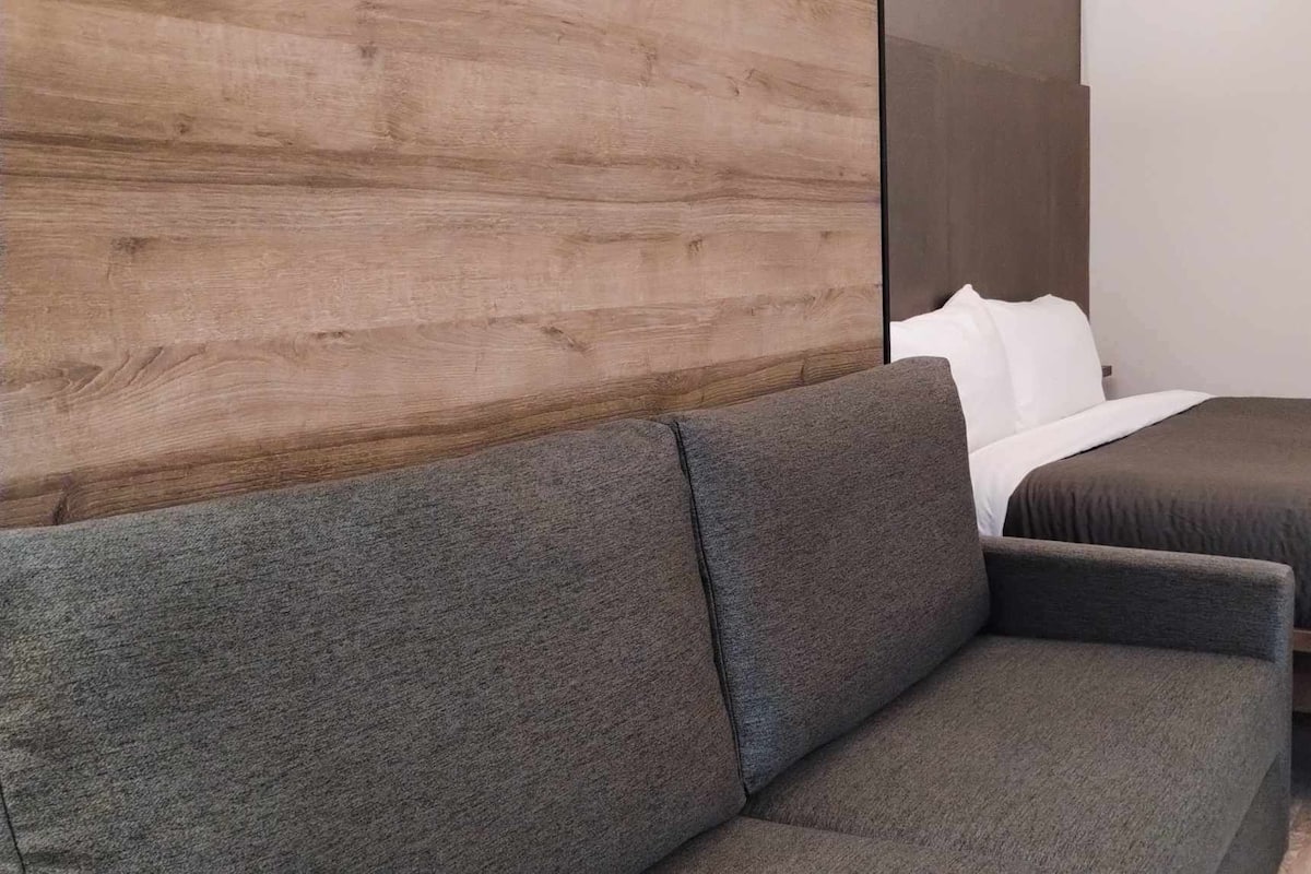 Une chambre d’hôtel moderne avec un canapé gris, un lit avec une literie blanche et un mur décoratif en bois.