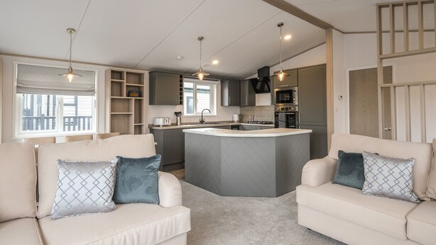 Een moderne keuken met een centraal eiland, inbouwkeuken en hanglampen. In een woonruimte staan twee banken met gekleurde kussens.