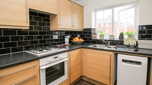 A modern kitchen with black and white tiles, wooden cabinets, a built-in oven, a dishwasher, and a window with a view of buildings. - 30 Poplar Close (Shrewsbury)