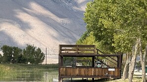 A wooden deck with a rustic design overlooking a calm lake, surrounded by lush greenery and a mountainous backdrop.